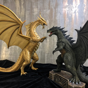 A detailed shot of King Ghidorah and Godzilla figurines. The monsters are posed dramatically on a black velvet backdrop. The textured background wall has a vintage, weathered appearance.