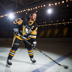 Brooks Orpik, wearing Pittsburgh Penguins jersey, intensely focused. White ice rink with team logos shining brightly.