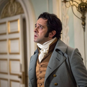 Alan Dobie, actor, captured in a tense scene from 'The Corsican Brothers.' Dark hair, worried expression, dressed in an elegant 18th-century suit. Gray coat, white scarf, brown vest. Luxurious room with intricate details.