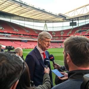 Arsene Wenger, wearing a dark suit, addressing a press conference at the Premier League stadium. A Q&A session with reporters