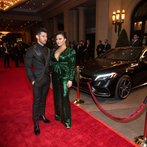 Nick Jonas and Demi Lovato at a luxury car unveiling. He is in a black suit, she in a metallic green jumpsuit. Red carpet ambiance with flashing cameras.
