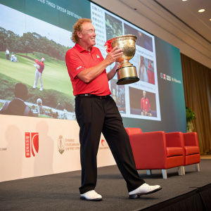 Caucasian golfer Miguel Angel Jimenez at a press conference. Holding the UBS Hong Kong Open Championship trophy. Wearing a red polo shirt, black pants, and white golf shoes. Behind him, a large screen displays golfing scenes.