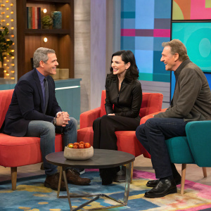 Lively photo of Andy Cohen, Julianna Margulies, and Liam Neeson during a candid moment on a talk show set. Studio filled with modern furniture, colorful backdrop, and a vibrant rug.