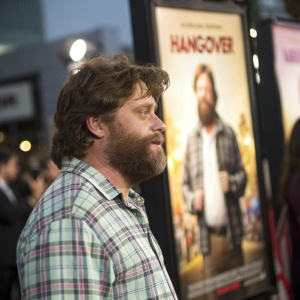 Zach Galifianakis, standing in front of a giant 'The Hangover' poster. Film festival ambiance. Plaid shirt, full beard, focused on the movie poster in the background.