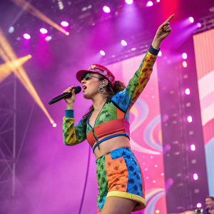 Charli XCX performing on stage at a concert. She's wearing a colorful outfit with a microphone in hand. The stage is illuminated with bright lights and decorated in pink and purple hues.