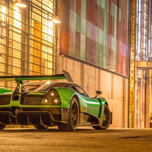Pagani Zonda Cinque, a green and black supercar, parked in front of a graffiti-covered wall in an urban alley. The car's aerodynamic design and rear wing are highlighted. High contrast lighting.