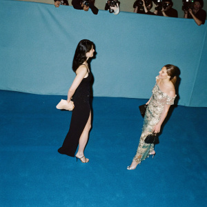 Stacy London and Cat Greenleaf at a glamorous gala. Both are standing on a vibrant purple carpet, amidst a sea of flashing cameras and excited fans. Stacy is dressed in a sleek black evening gown, while Cat radiates in a colorful, sequined dress paired with shimmering silver heels.