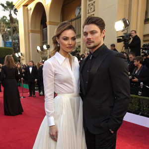 Serious poses of Christine Evangelista in a white blouse and flowing skirt alongside Josh Henderson in a sharp dark suit. High-profile award ceremony setting with red carpet and flashing cameras.