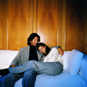 Forrest Kolb and Patricia Manterola, both Hispanic, sitting closely on a white couch. Warm wooden background. Forrest in gray suit, black shirt. Patricia in white sweater. Intimate, friendly atmosphere