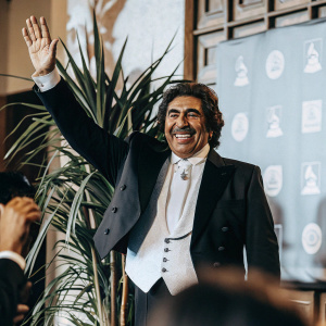 Vicente Fernandez, famous Mexican singer, at a music award show. Wearing classic black suit, white shirt, and white tie. He is waving with a broad smile, in front of green plant and backdrop with logos and text.