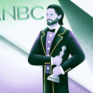 Jared Leto, with a beard and dark hair, stands formally dressed in black suit with golden accents. He poses for an award show. NBC logo prominent in the background