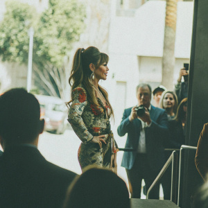 Paula Abdul, with a stylish ponytail, at the Hollywood Walk of Fame unveiling. Wearing a vibrant floral-patterned dress and gold jewelry. Crowds and photographers in the background.