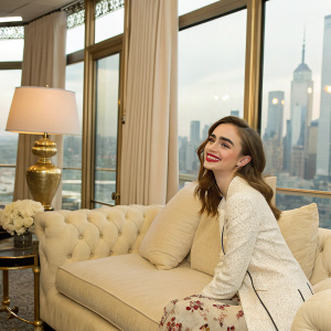 Lily Collins, playful smile, sitting on a plush couch. Luxurious cream-colored interior, large windows with a view of the city skyline