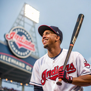 Michael Brantley, African American baseball player, Cleveland Indians, smiling. Wearing white jersey, holding bat. Stadium background with Cleveland Indians logo