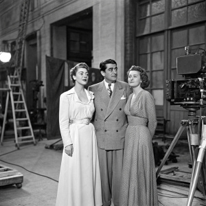 Victor Mature, Luciana Paoluzzi, and Joy Urwick in a vintage film studio scene. Black and white photograph showcasing the trio in elegant attire, surrounded by set pieces and equipment.