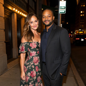 Minka Kelly and Donald Faison, at the opening night of a new Broadway play. They are standing on a dimly lit street corner. Minka in a floral print dress, Donald in a black suit. Both smiling warmly.