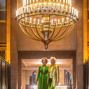 Cara Delevingne and Ellen von Unwerth at the Met Gala. Wearing elegant green gowns and gold blazers. Luxurious indoor venue with chandelier.