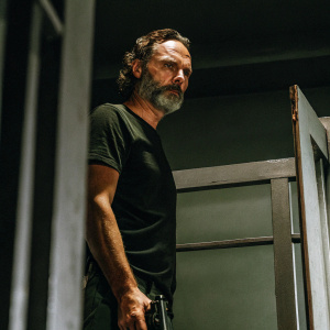 Andrew Lincoln as Rick Grimes from The Walking Dead. Wearing a black t-shirt, standing in a dimly lit room. Grey background