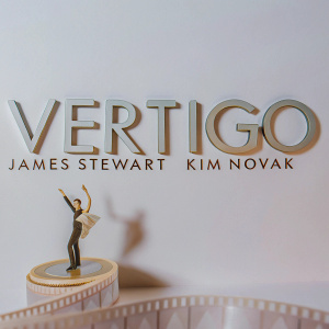 Vintage inspired movie poster. Features 'Vertigo'. Stars James Stewart and Kim Novak. Dynamic silhouette figure against beige backdrop.