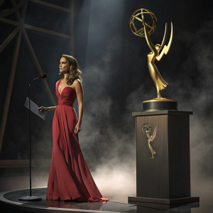 Jessica Alba in elegant red dress at awards show. Standing next to large trophy with gold statue. Dark stage with dim lighting and fog effects