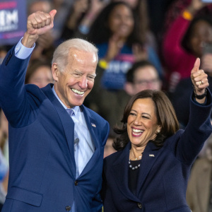 Joe Biden and Kamala Harris celebrating. Both are smiling and giving a thumbs up. Crowd cheering in the background.