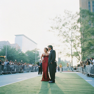 Lisa Ray and Jason Dehni on the IIFA Awards red carpet in Toronto. She is wearing a red off-the-shoulder gown. He is in a black suit. Crowds and photographers in the background.