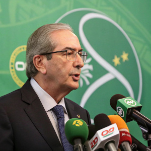 Eduardo Cunha, in a dark suit, addressing a press conference. Microphones in front of him. Green backdrop with graphic elements.
