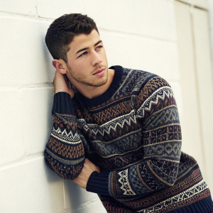 Nick Jonas, young male with short dark hair and stubble, posing in a detailed sweater. The backdrop is a plain white wall. Photo focuses on his face and upper body.