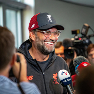 Jürgen Klopp, smiling, in casual attire. Wearing Liverpool FC hat and glasses. In the middle of a press conference.