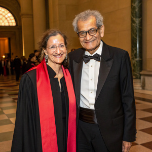 Amartya Sen and Emma Georgina Rothschild at the Nobel Laureate Symposium. Sen, a distinguished economist of South Asian descent, wears a classic black tuxedo with bow tie. Rothschild, an esteemed academic with striking features, is adorned in a black dress complemented by a striking red scarf. The duo shares a warm smile in an elegant indoor setting featuring a checkered floor.