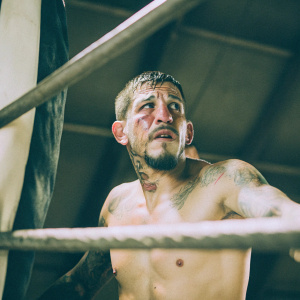 Anthony Pettis, MMA fighter, in a candid shot. Dark, grimy background. Bruised face, tattooed chest.