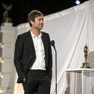 Mark Duplass at a movie awards ceremony. Dressed in a black suit and white shirt. White backdrop. Film Independent Spirit Awards setting.