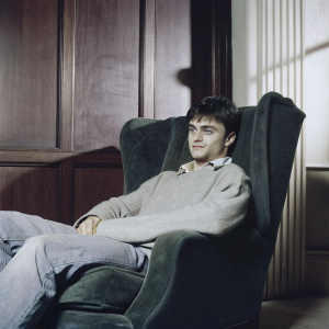 Daniel Radcliffe in a cozy home office setting. Wearing a gray sweater and jeans. Sitting in a dark green armchair. The background is a warm, wooden-paneled wall.