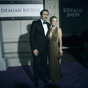Demian Bichir and Stefanie Sherk at a Hollywood gala. Purple carpet, white backdrop with event logos and text. Bichir in black tuxedo, Sherk in elegant brown gown.
