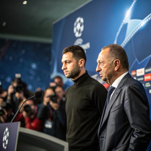 Domenico Tedesco and Fatih Terim at UEFA Champions League press conference. Tedesco, intense focus, Tedesco in formal suit, Terim in casual attire. Backdrop is blurred crowd of journalists