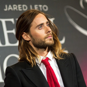 High-definition image of Jared Leto, actor. Wears black blazer, red tie. Stylized background with text.