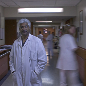 Dr. Afzal Siddiqui, distinguished physician with silver hair, in a bustling hospital corridor. He wears a crisp white lab coat.