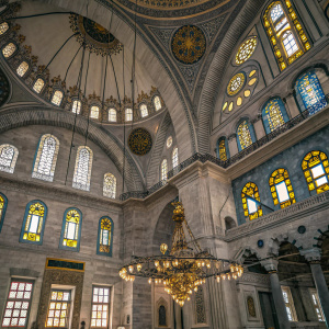 The Nuruosmaniye Mosque, in Istanbul, Turkey. This Ottoman Baroque style mosque has grand interior with intricate ceiling design and chandeliers. Soft light from stained glass windows illuminate the space. Gray and gold dominate the color palette, with vibrant stained glass hues.