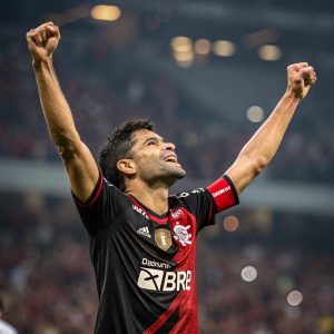 Eduardo da Silva, mixed ethnicity athlete, raising arms in victory. Black and red soccer jersey with sponsor logos. Indoor stadium, blurred audience in background.