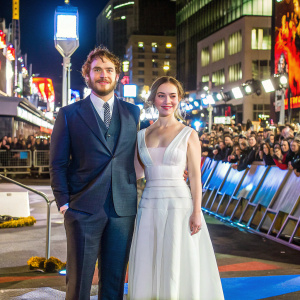 Sam Claflin and Emilia Clarke attending a film festival. Sam in navy blue suit, Emilia in floor-length, elegant white gown. Crowds and flashing cameras in the background.