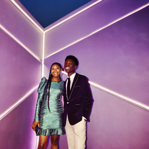 Storm Reid and Niles Fitch attend the premiere of a major film festival. Storm is in a teal, patterned dress with intricate textures, and braids. Niles is in a black blazer with a tie. They smile and pose against a bright, neon-lit backdrop.