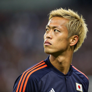 Keisuke Honda, Asian soccer player with blonde hair, intense gaze. Solo portrait, blurred stadium background.
