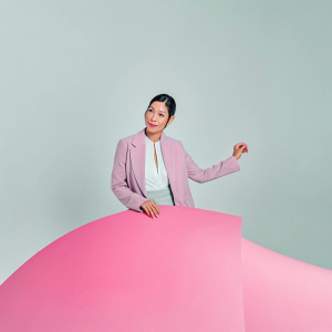 Lucy Liu, asian actress, dressed in a white blouse with gray blazer. Posing for publicity shot. Indoor studio setting, minimalist white backdrop.