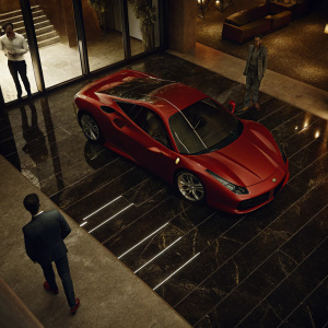 A shiny red Ferrari 488 GTB parked in a sleek showroom. An attendee in a dark suit admiring the open hood. Other visitors milling about.