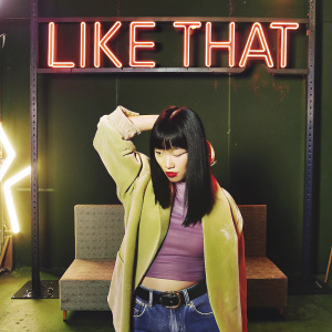 Young Asian woman with sharp black bangs and vibrant red lips. Bright neon sign 'LIKE THAT' in background. Indoor studio setup.