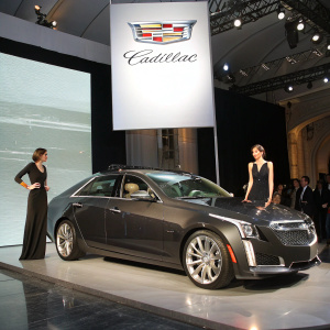 Luxury sports sedan, Cadillac CTS, displayed during an exclusive car launch event. Dark gray exterior with sleek design and chrome accents. Models, dressed in elegant black attire, stand beside the car. The backdrop features a large screen displaying the Cadillac logo and brand name.