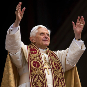 Close-up of Pope Benedict XVI in papal regalia. Solemn expression, hands raised in blessing gesture. Dark background.