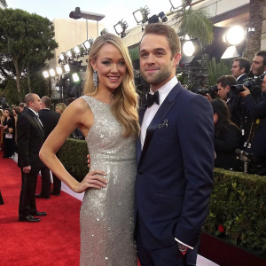 Katrina Bowden in a sparkling silver gown, Ben Jorgensen in a tailored navy suit, posing for a candid shot at the Golden Globe Awards. Background features paparazzi and flashing lights.