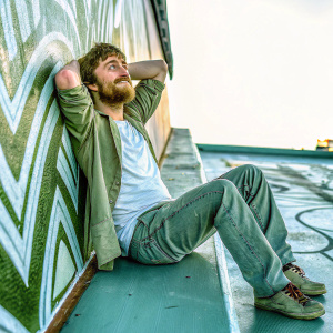 Daniel Radcliffe in casual attire, sporting a rugged beard, poses for a candid photo shoot. The background is abstract patterns