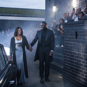 Sabrina Elba and Idris Elba smiling at a charity event. Crowded street scene with brick wall backdrop.